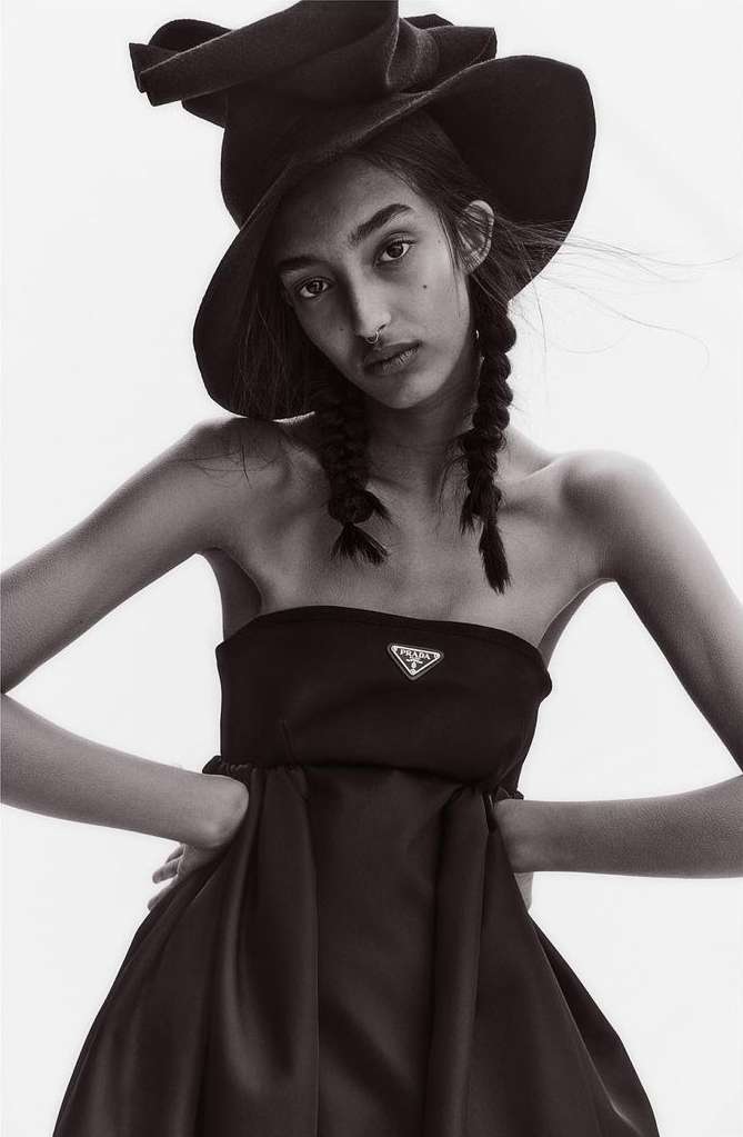 Model with braided hair wearing a black strapless dress and wide-brimmed hat against a plain background