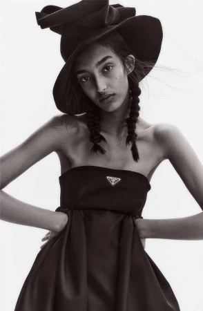 Model with braided hair wearing a black strapless dress and wide-brimmed hat against a plain background