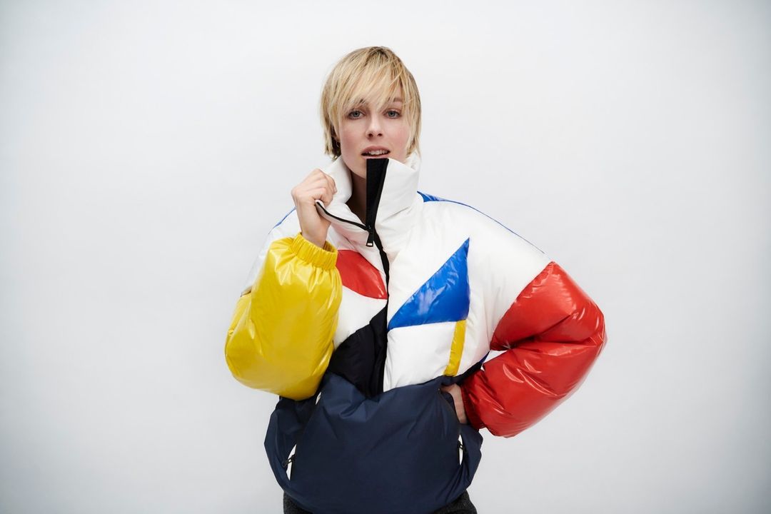 Adult woman with blonde hair in a color-blocked puffer jacket with yellow, red, blue, and white against a plain background.