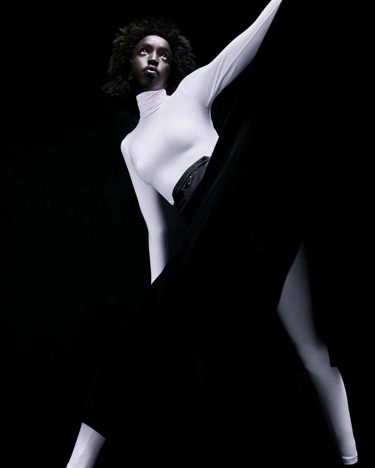 Model in white turtleneck and black skirt posing against a dark background in a high-contrast portrait captured by Bibi Cornejo Borthwick.