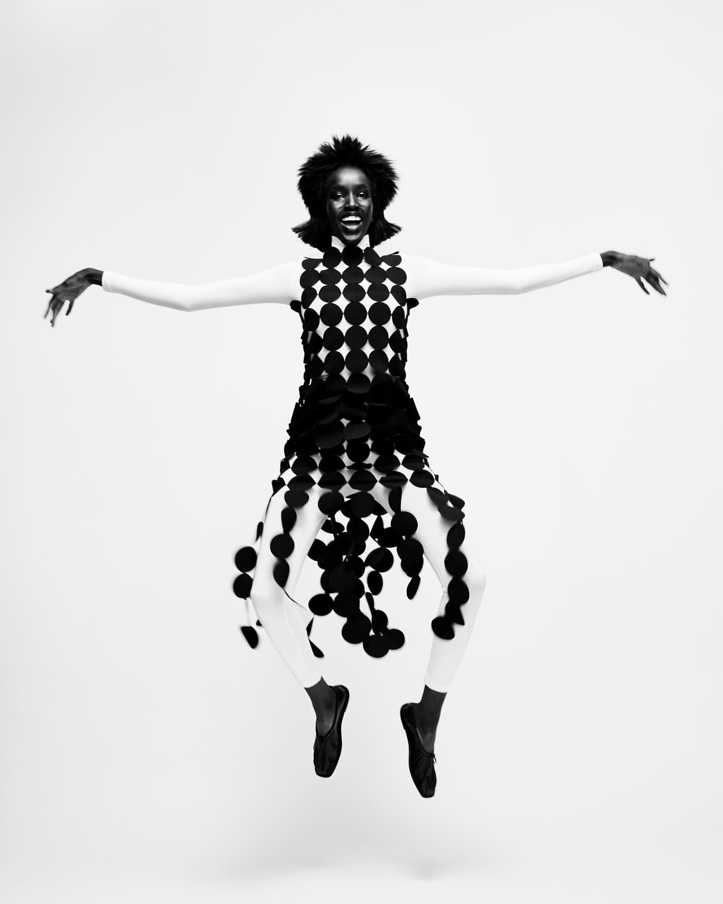 A woman wearing a polka dot dress is jumping in a studio. The model is wearing full body stocking and has her arms spread, black and white photo.