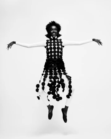 A woman wearing a polka dot dress is jumping in a studio. The model is wearing full body stocking and has her arms spread, black and white photo.