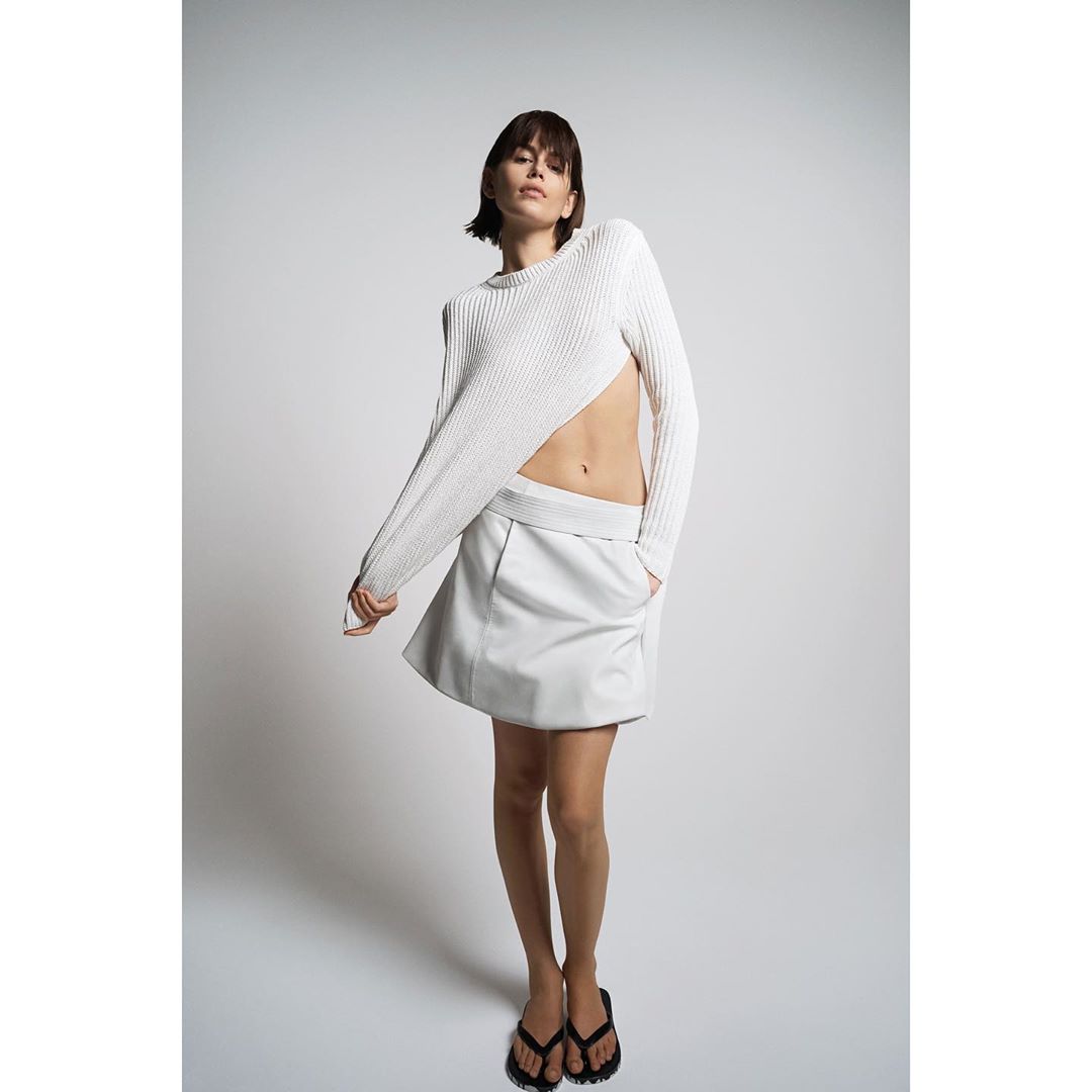 A woman poses in a studio wearing a white, long-sleeved ribbed sweater, a white leather miniskirt, and black flip-flops in a fashion shot.