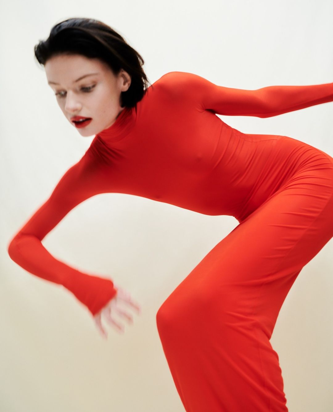 Woman in a full red bodysuit with long sleeves striking a dynamic pose against a neutral backdrop, showcasing fashion and movement.