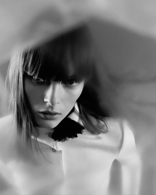 Monochrome fashion portrait of a young woman with dark hair, wearing a light-colored top with a frilled collar, looking down.