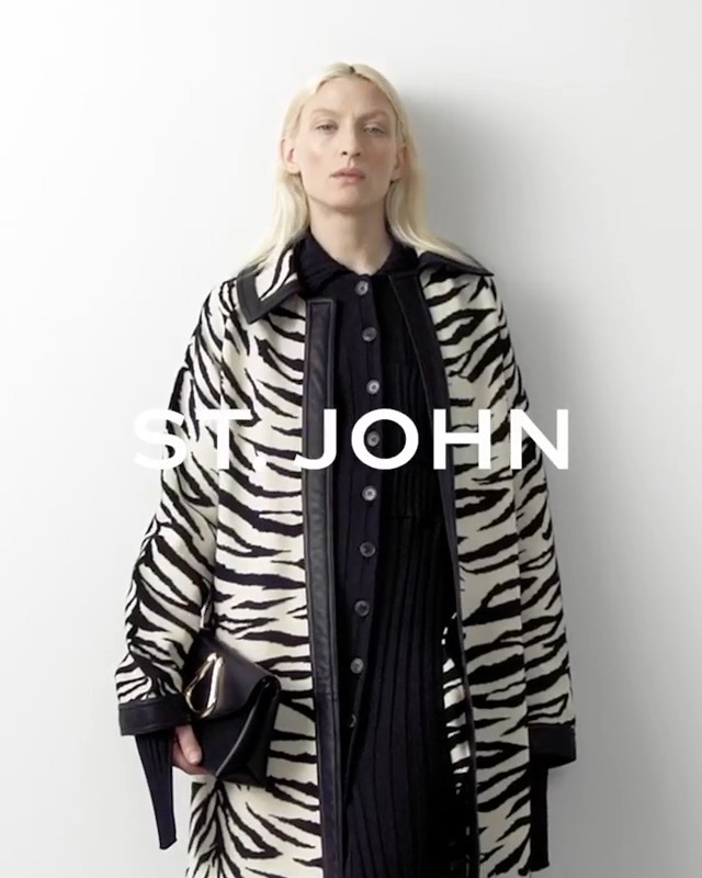 A blonde adult female model showcases a black and white zebra-striped coat, a black dress, and a black purse against a plain white background.