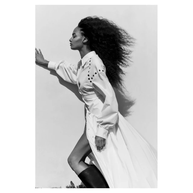 Elegant woman in a white dress and black boots leans against a white wall; wind blows through her curly, dark hair in black and white.
