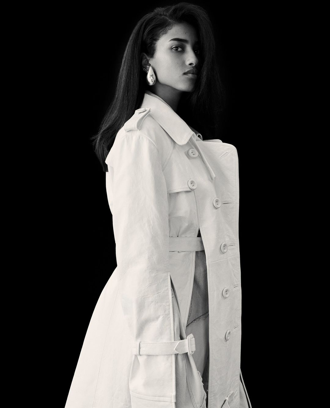 Black and white portrait of a stylish adult female wearing a light colored trench coat and elegant earrings against a dark background.