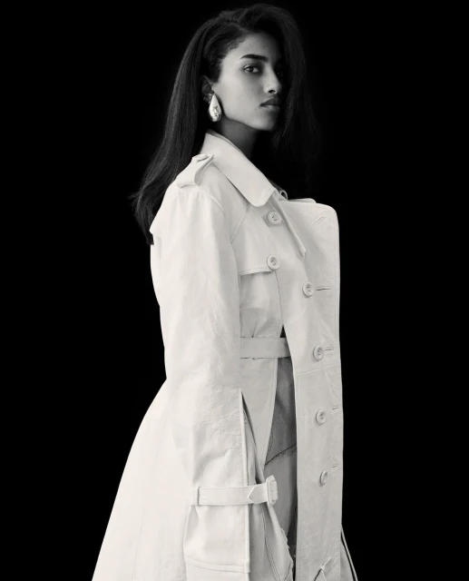 Black and white portrait of a stylish adult female wearing a light colored trench coat and elegant earrings against a dark background.