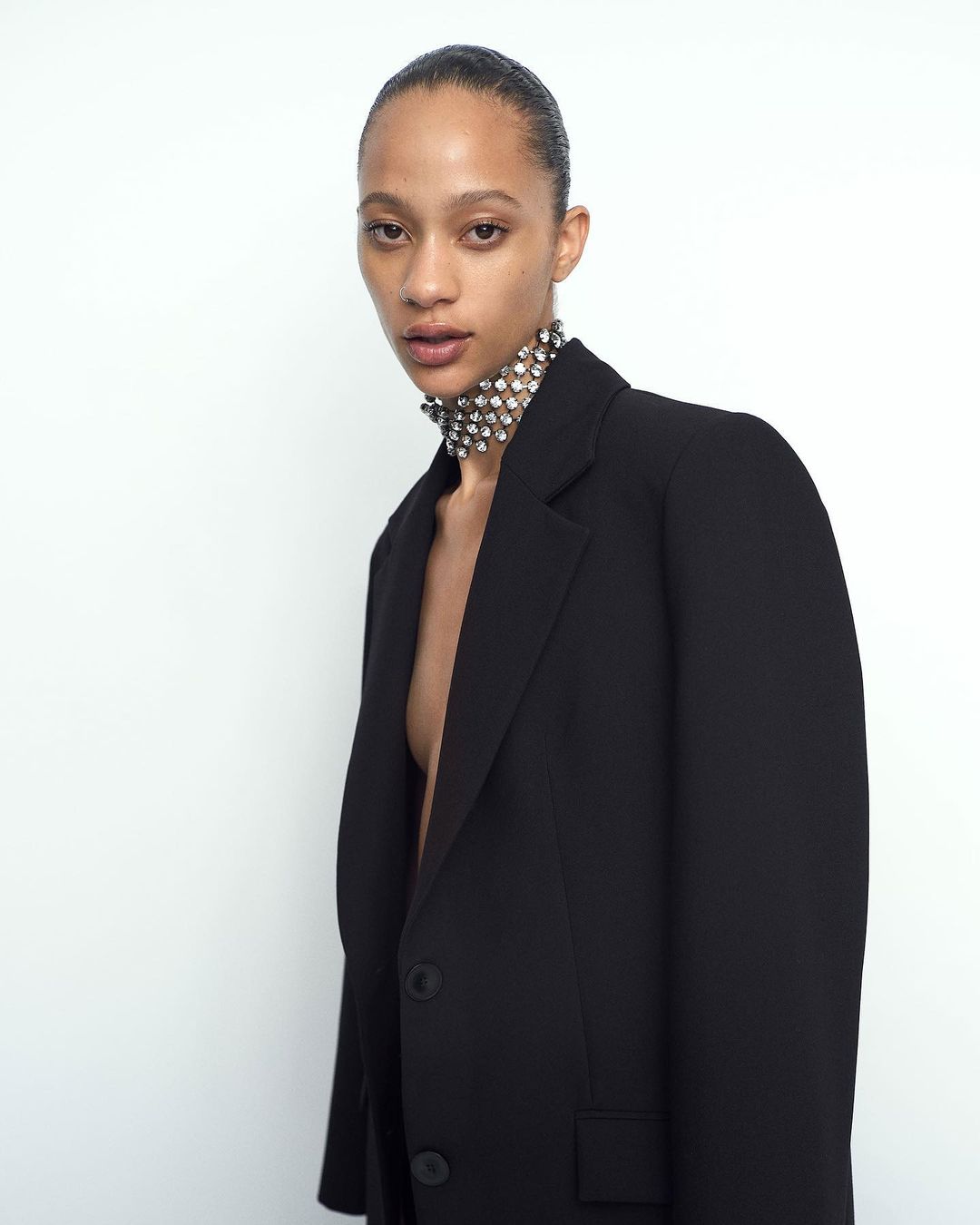 Fashion shot of a black female model wearing a black blazer and crystal choker against a white background in a studio setting.