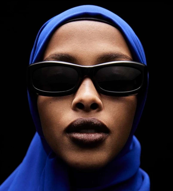 Close-up portrait of a woman with dark skin wearing black sunglasses and a blue headscarf against a stark black background.