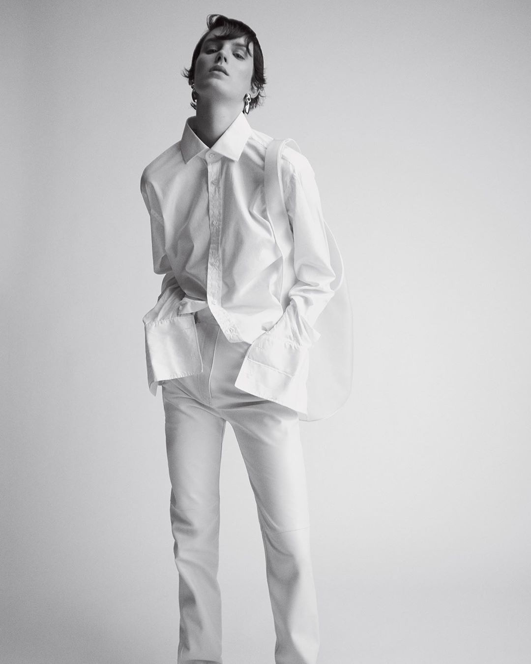 Studio shot of an adult woman in a white collared shirt and white pants, posing against a light gray seamless backdrop.