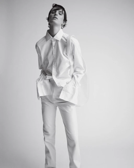 Studio shot of an adult woman in a white collared shirt and white pants, posing against a light gray seamless backdrop.