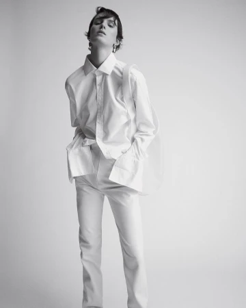 Studio shot of an adult woman in a white collared shirt and white pants, posing against a light gray seamless backdrop.