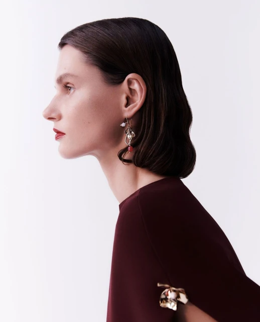 Elegant adult woman in a side-profile headshot wearing a burgundy top and striking jewelry against a clean white background.