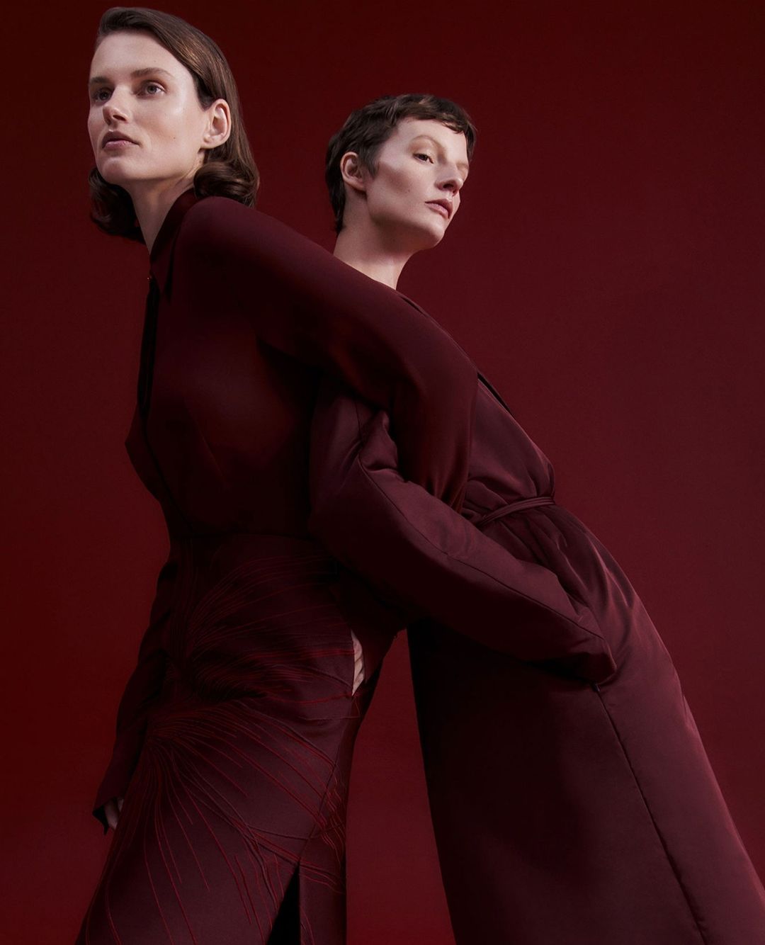 Two women stand back to back in a studio setting, wearing dark red dresses against a matching dark red backdrop. Fashion editorial shot.