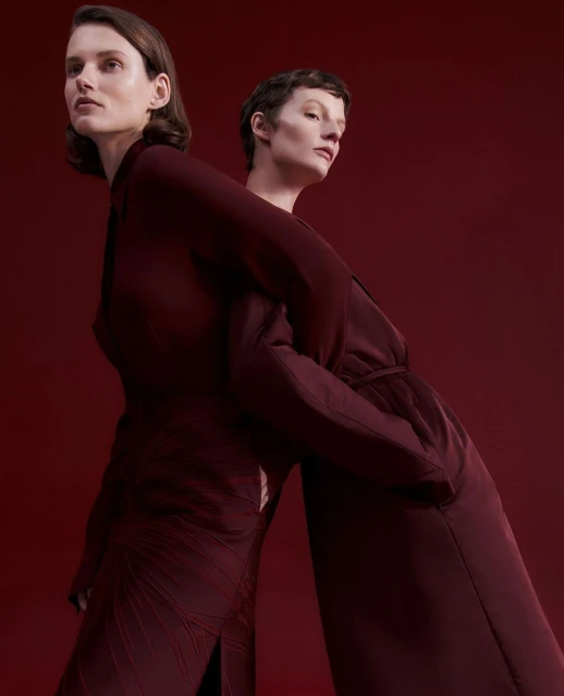 Two women stand back to back in a studio setting, wearing dark red dresses against a matching dark red backdrop. Fashion editorial shot.