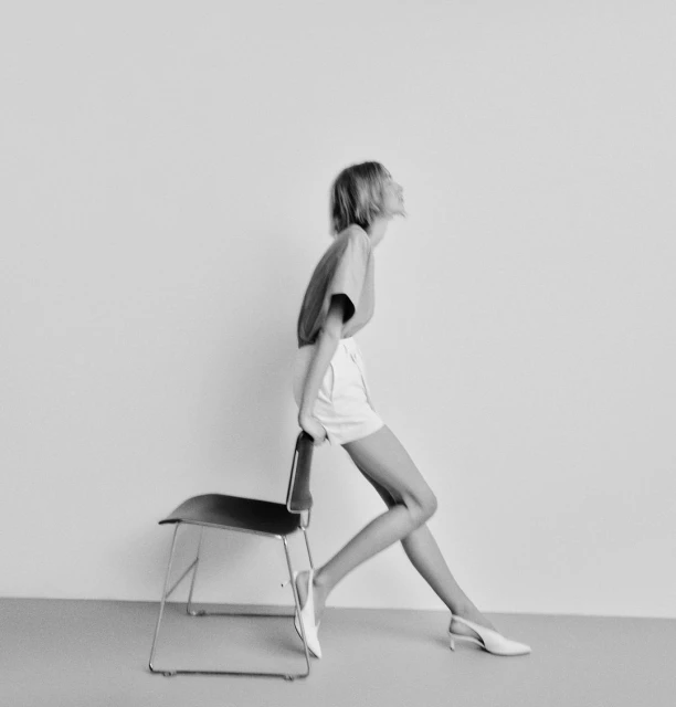 Woman in t-shirt and shorts leaning on a chair in a minimalist studio setting, creating a modern and stylish black and white image.