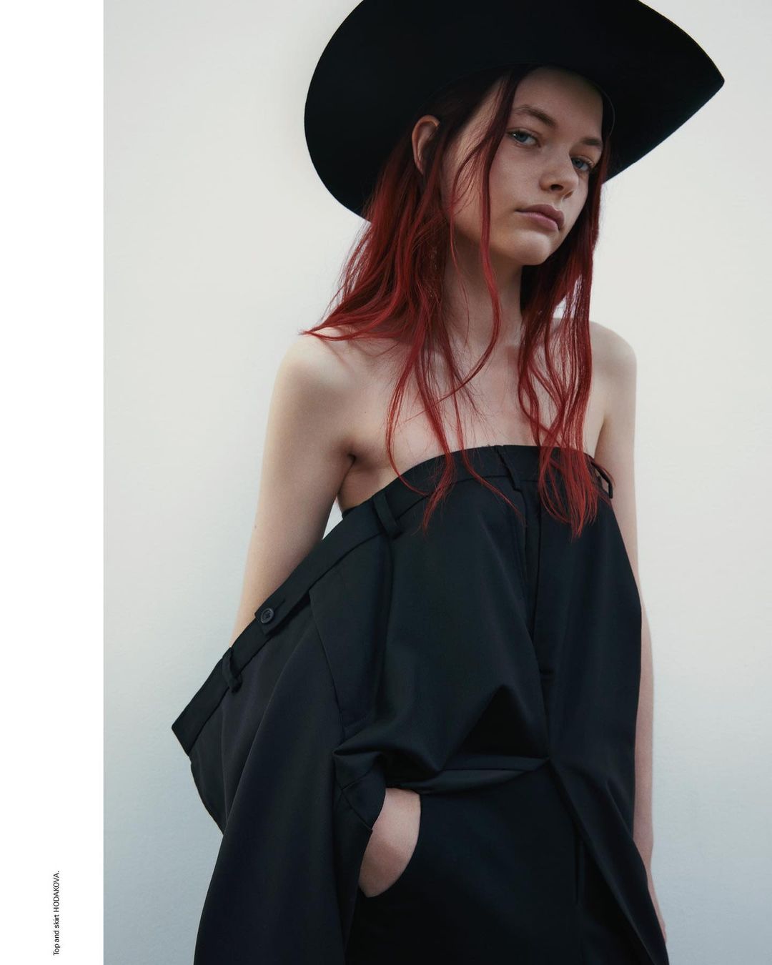 A portrait of a woman with red hair wearing a wide black hat and a strapless black garment against a plain white background.