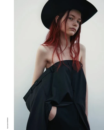 A portrait of a woman with red hair wearing a wide black hat and a strapless black garment against a plain white background.