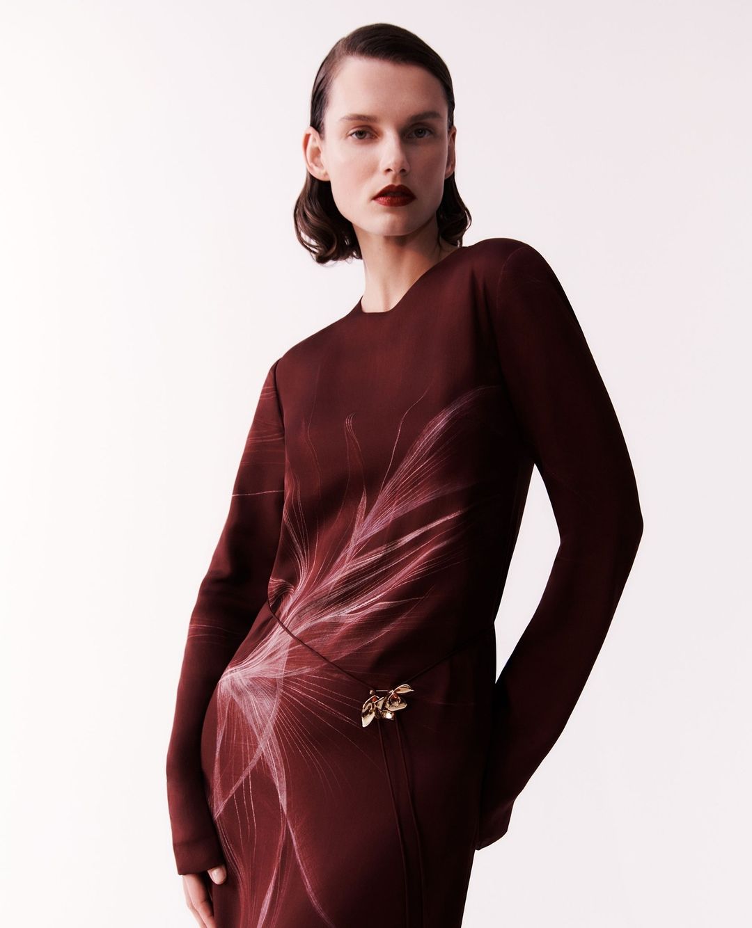 Adult female fashion model in a long-sleeve burgundy dress with a floral pattern posing in a photo studio setting on a seamless background.