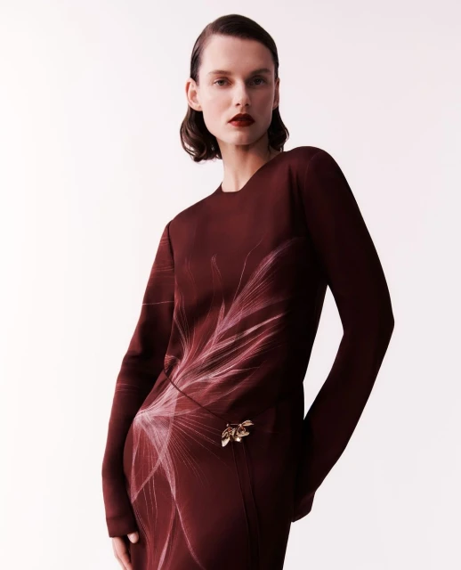Adult female fashion model in a long-sleeve burgundy dress with a floral pattern posing in a photo studio setting on a seamless background.