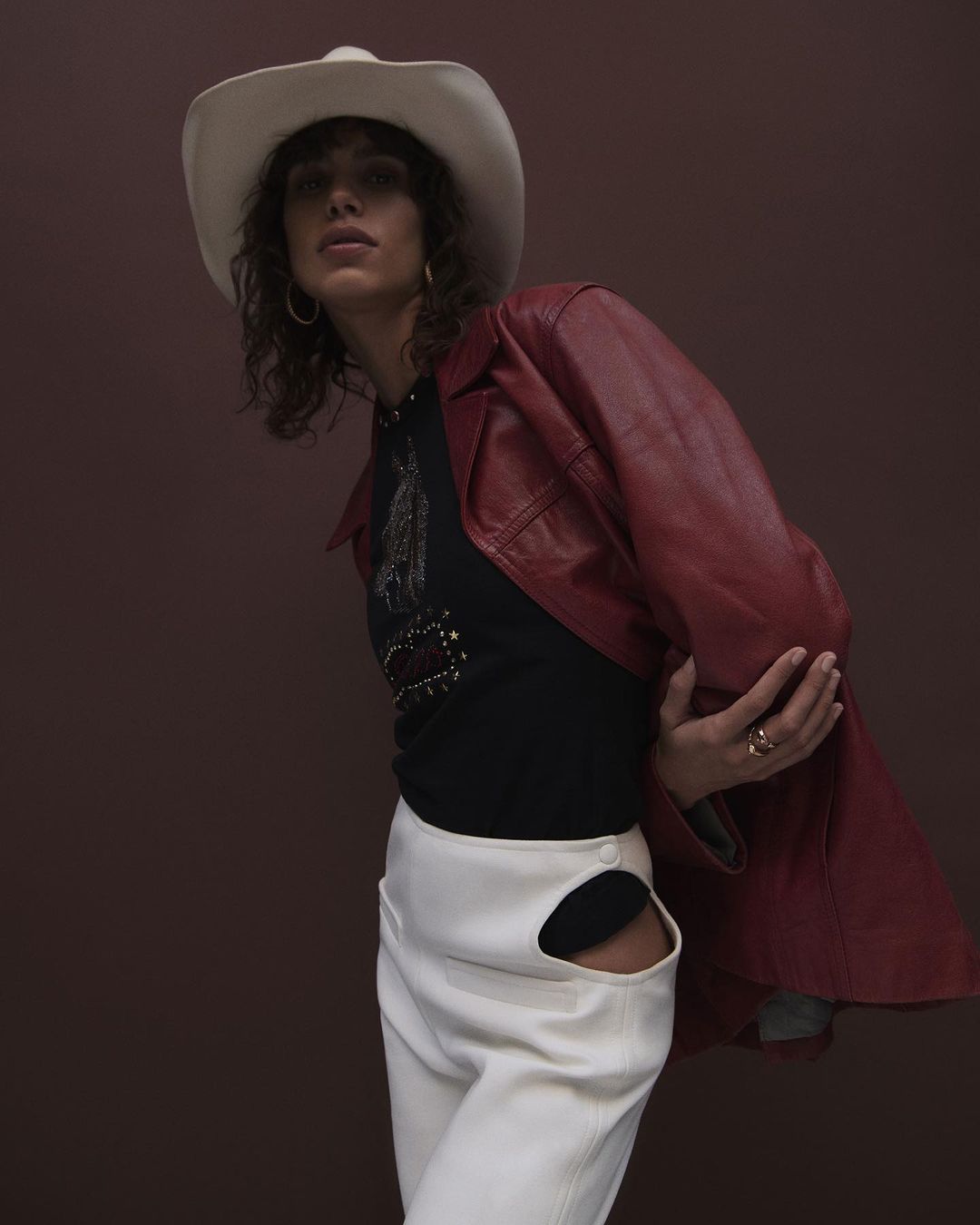 Portrait of a model in a black top, white pants with cutouts, a red jacket, and a cowboy hat against a brown backdrop.