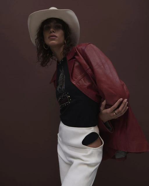 Portrait of a model in a black top, white pants with cutouts, a red jacket, and a cowboy hat against a brown backdrop.
