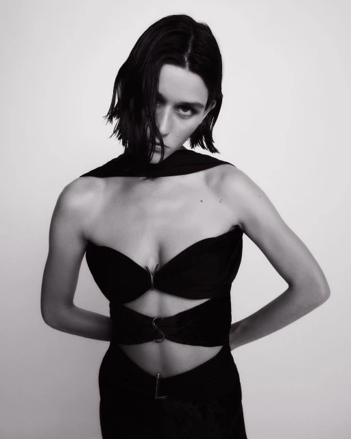 Black and white portrait of a woman in a stylish black dress against a neutral background, highlighting her dark hair and striking features.