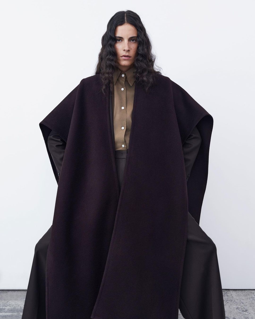 Studio shot of a woman in a brown button-down shirt and a plum-colored coat with oversized sleeves against a white backdrop.