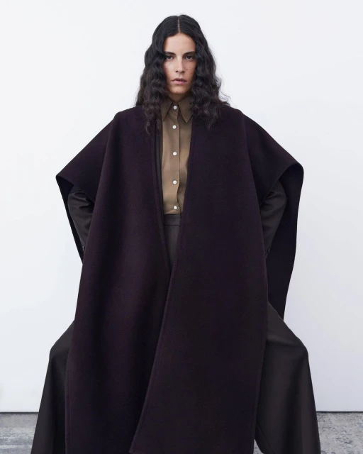 Studio shot of a woman in a brown button-down shirt and a plum-colored coat with oversized sleeves against a white backdrop.