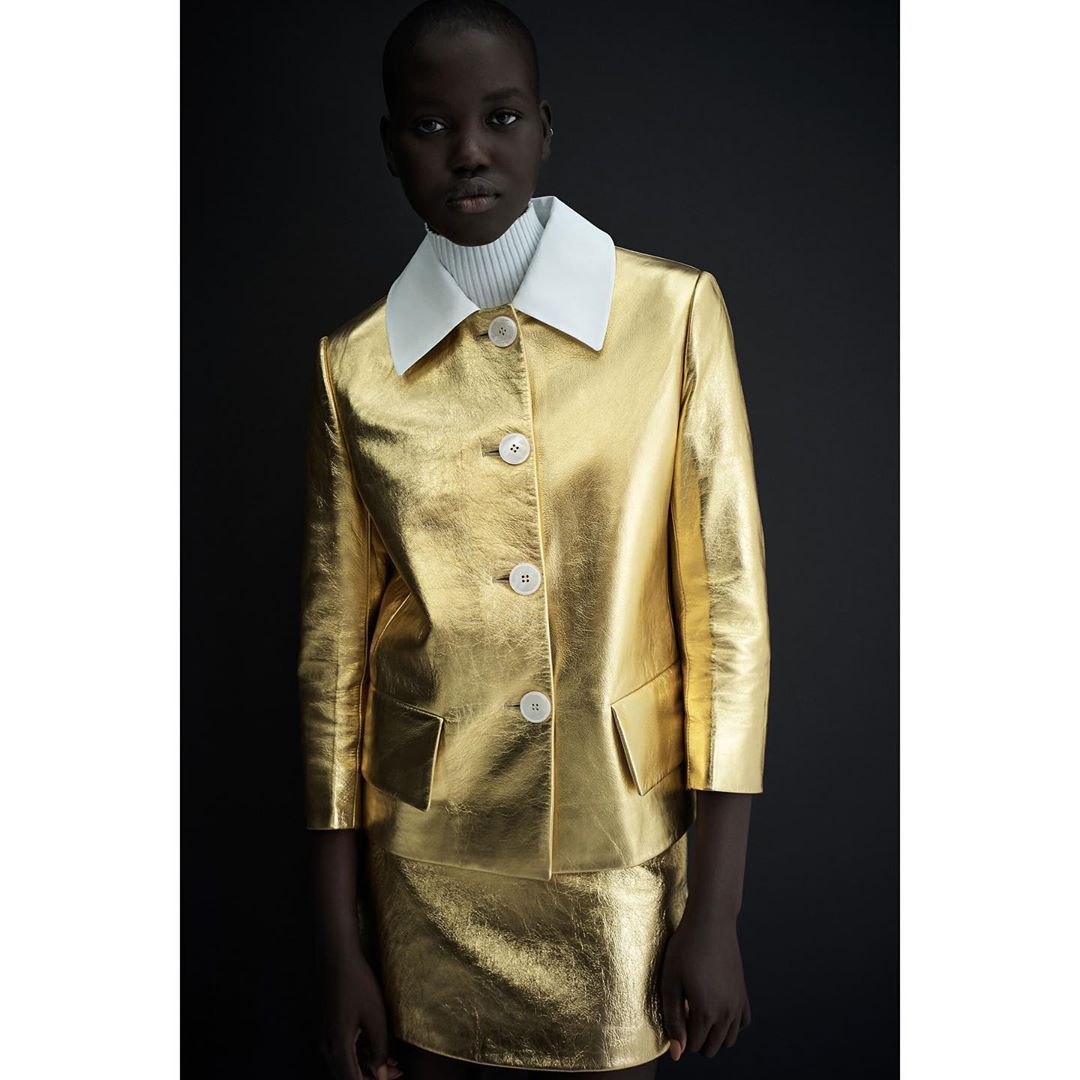An African-American woman presents in a studio setting, wearing a shimmering gold suit with a white-collared shirt, under dark, contrasting light.