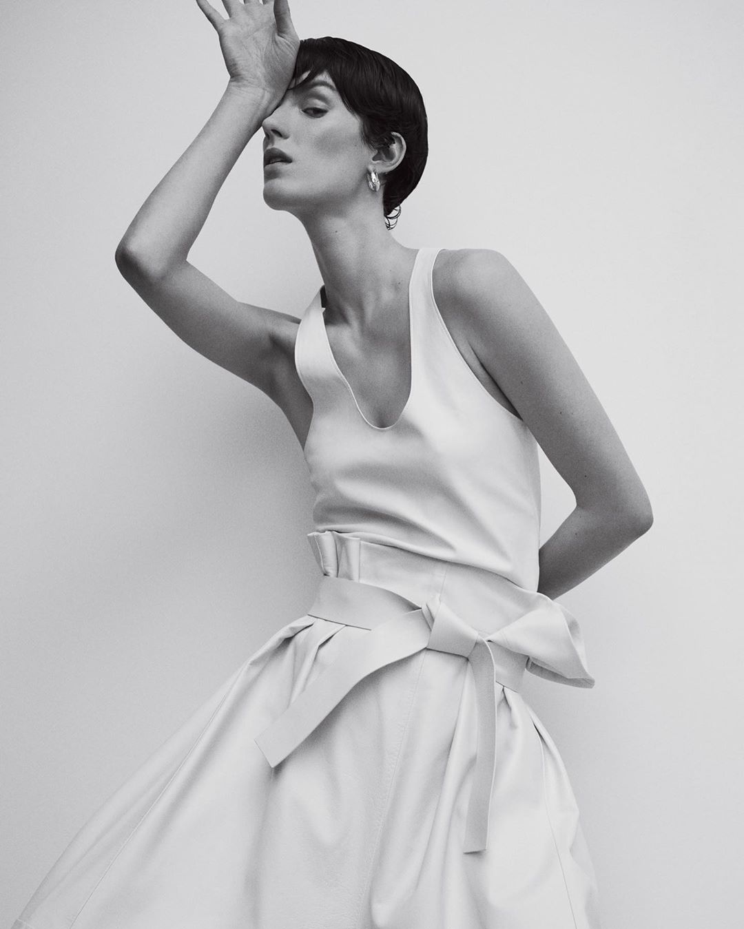 Model with short dark hair in white sleeveless top and pleated skirt posing with hand to forehead, photographed by Bibi Cornejo Borthwick.