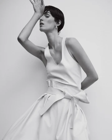 Model with short dark hair in white sleeveless top and pleated skirt posing with hand to forehead, photographed by Bibi Cornejo Borthwick.