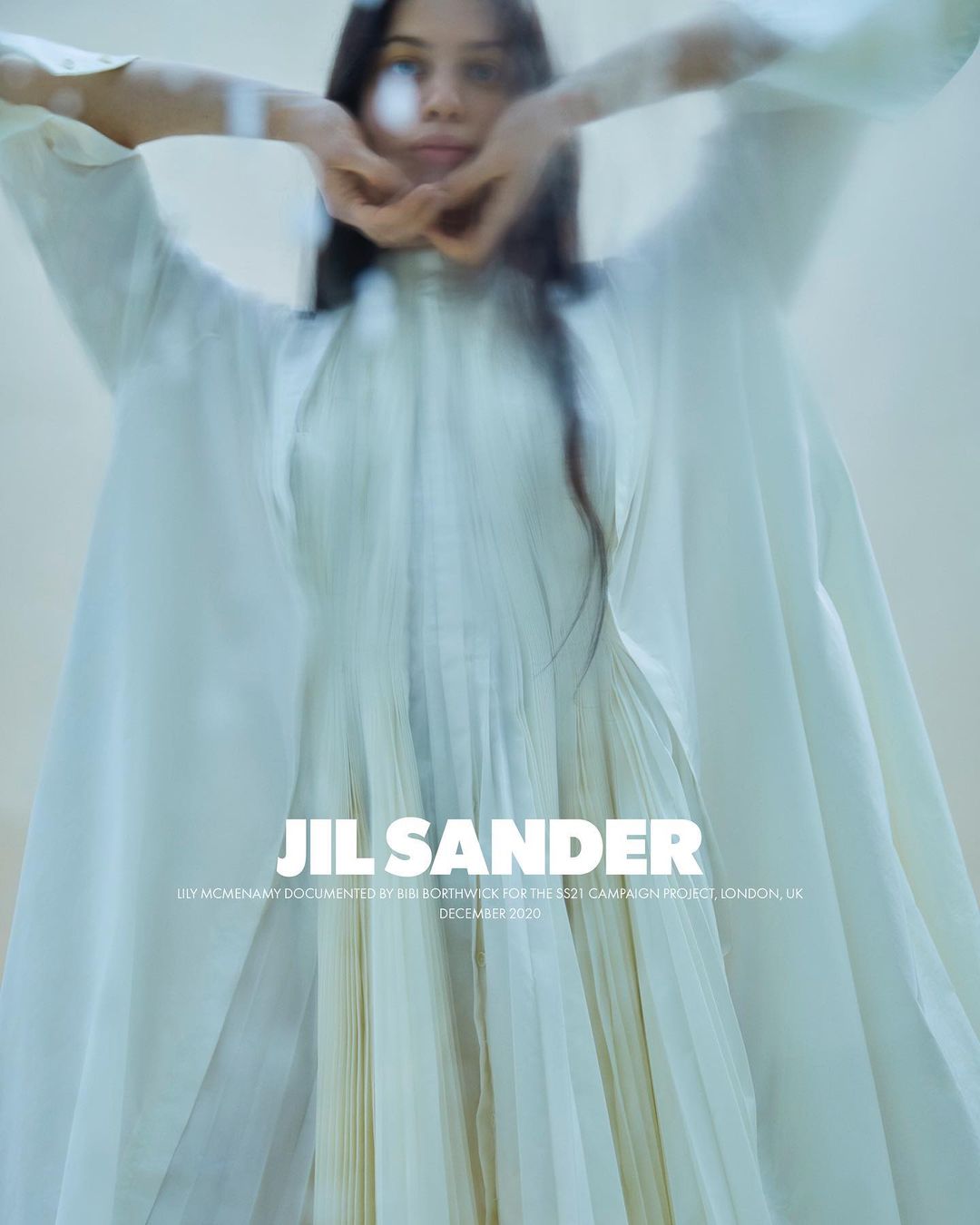 Fashion shot of a model in a flowing white gown, hands near her face. Jil Sander logo is visible, suggesting this is an ad campaign.
