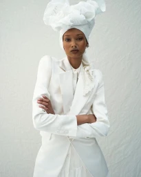 Model in white blazer with arms crossed wearing a matching white headwrap and septum ring against a textured backdrop