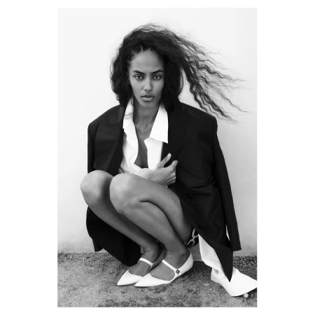 Black and white portrait of a woman crouching outdoors in a black blazer, white shirt, and white Mary Jane flats
