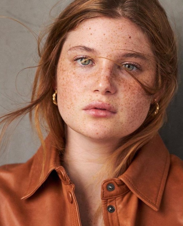 Close-up studio portrait of a red-haired woman with freckles wearing a tan leather shirt and gold hoop earrings against a neutral background.