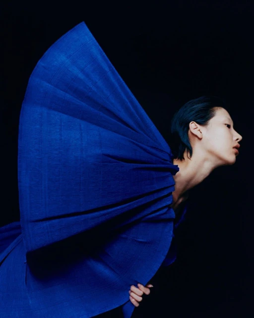 Portrait of an Asian woman wearing a draped blue garment against a dark background; fashion and beauty photography.