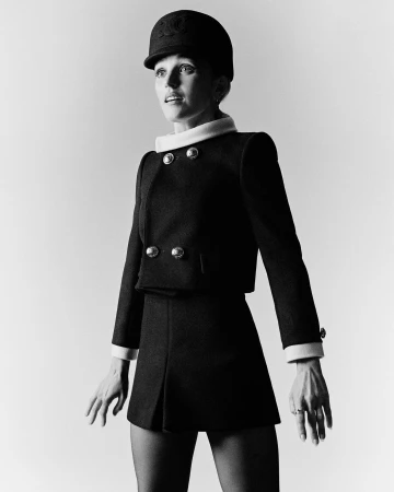 Model wearing a structured dark jacket, matching skirt, and jockey cap against a white background