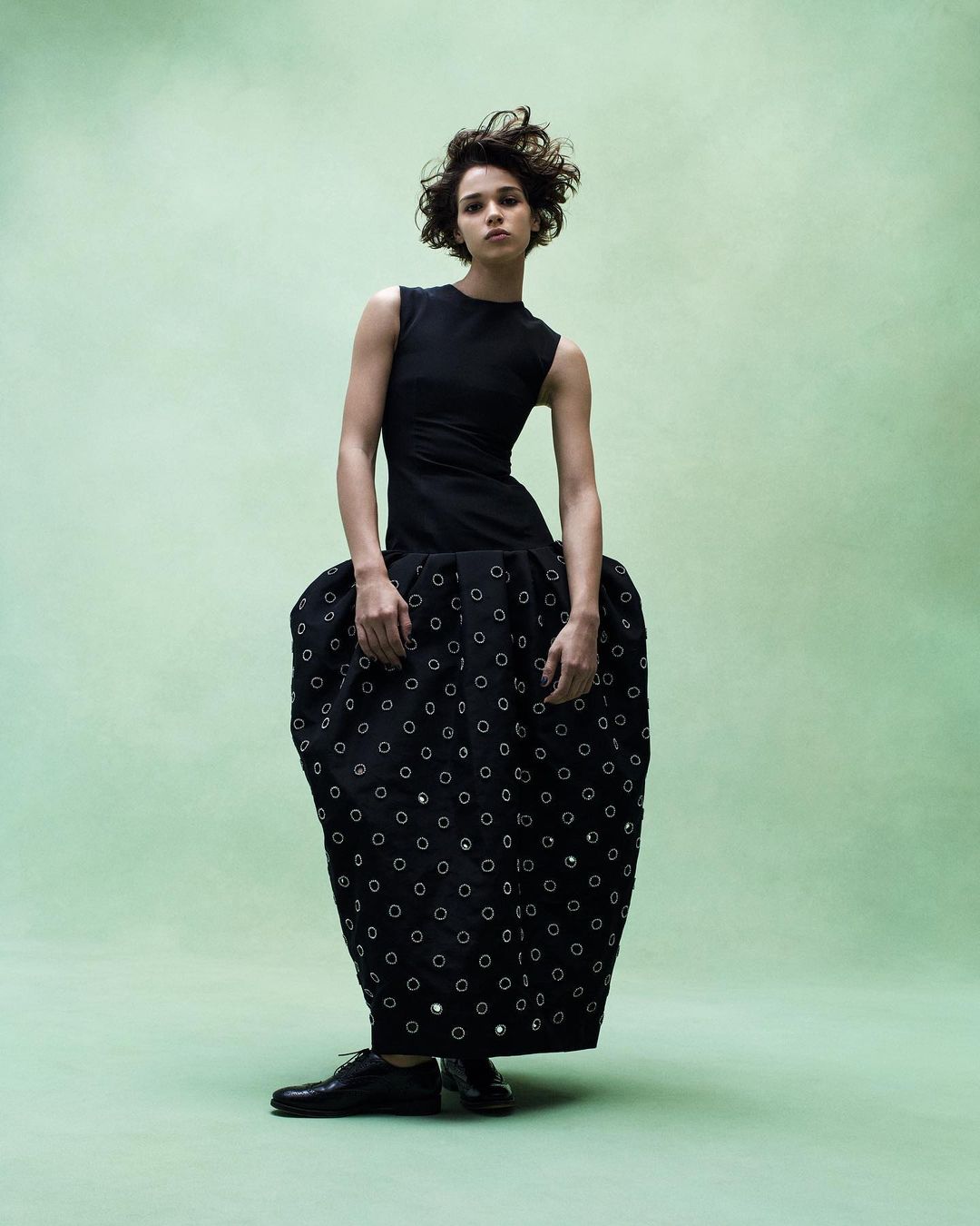 Fashion shot of a curly-haired woman posing in a black dress against a light green backdrop, showcasing unique design and style.
