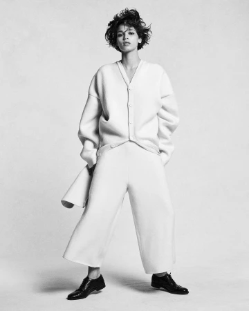Model with short curly hair wearing a buttoned cardigan and wide trousers against a light background