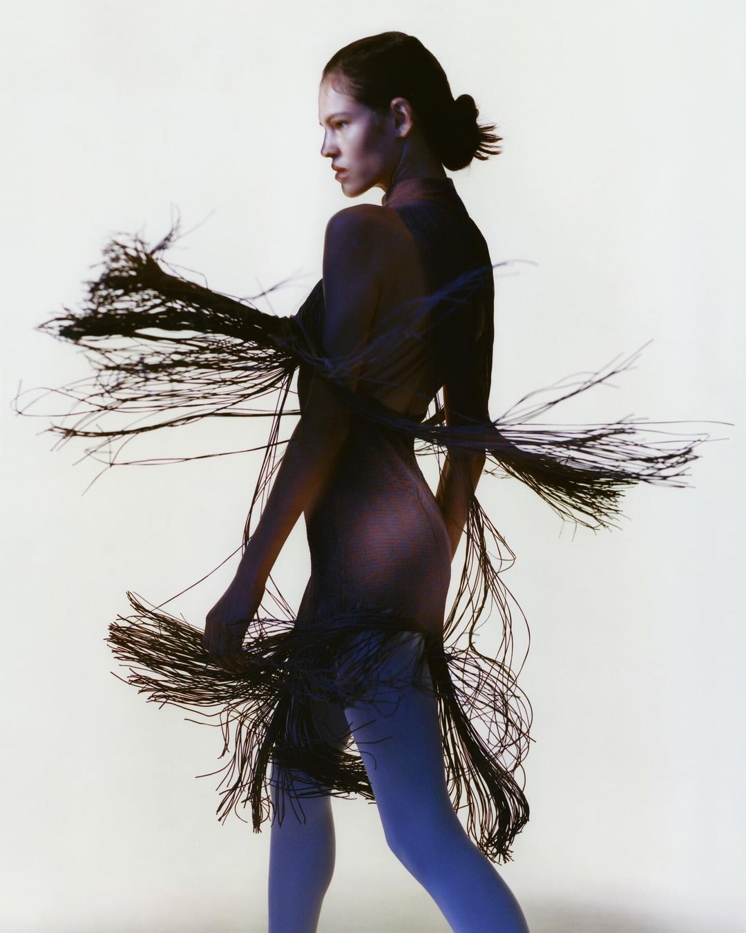 Woman in flowing black fringe dress, side profile in front of white backdrop. Studio lighting and fashion editorial aesthetic.