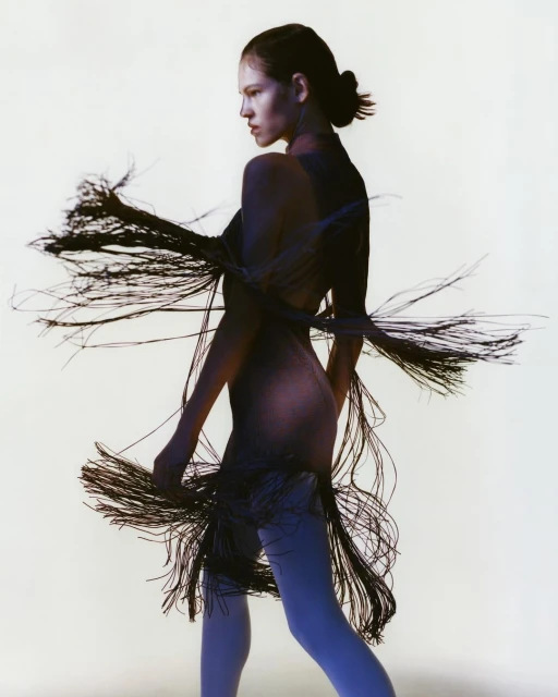 Woman in flowing black fringe dress, side profile in front of white backdrop. Studio lighting and fashion editorial aesthetic.