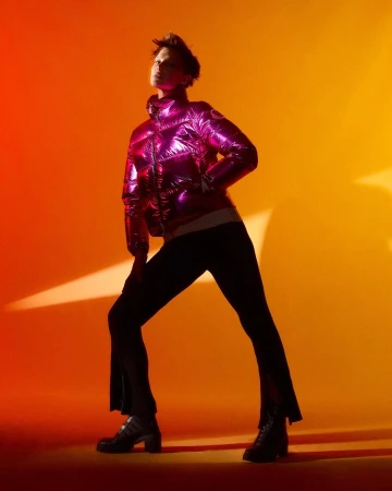 An adult female model strikes a full-length pose against an orange and yellow backdrop, wearing a pink puffer jacket, dark pants, and black boots.