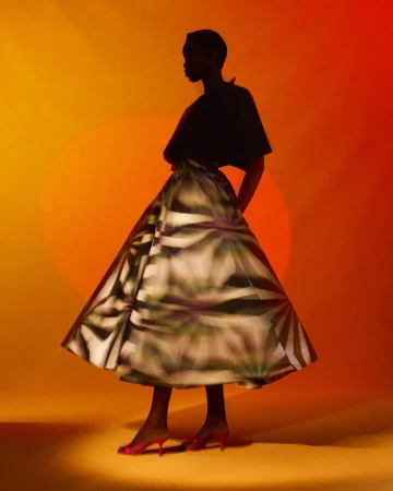 Fashion shot of a black, slim woman in a black top and patterned skirt against an orange background, standing and posing in pink heels.