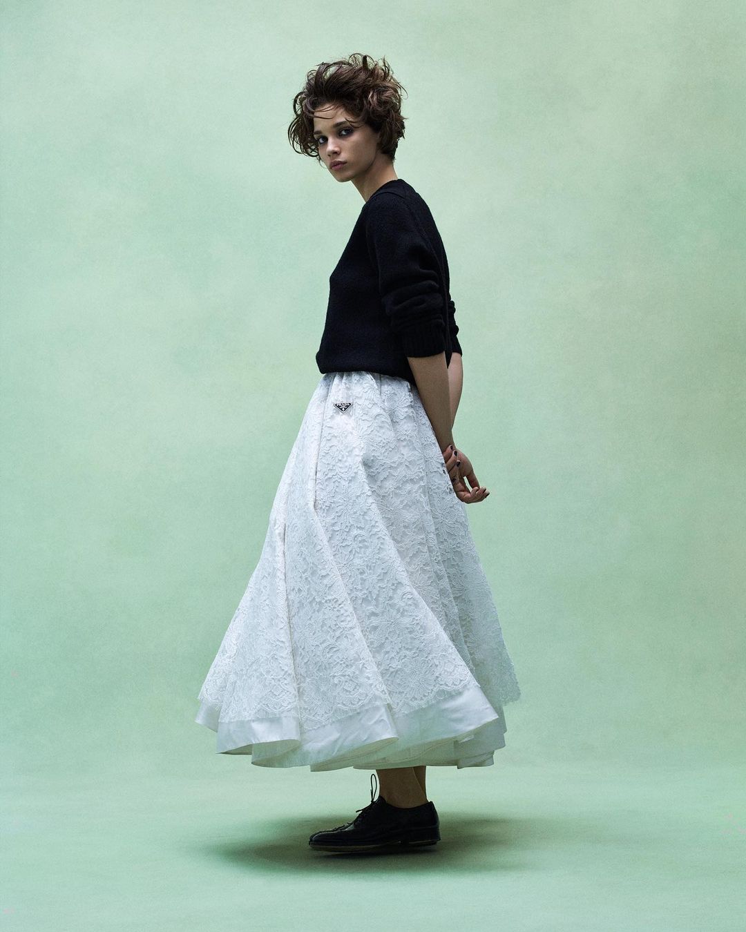 Model with short dark hair wearing a black sweater and white lace skirt against a soft green textured backdrop.