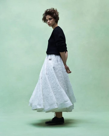 Model with short dark hair wearing a black sweater and white lace skirt against a soft green textured backdrop.