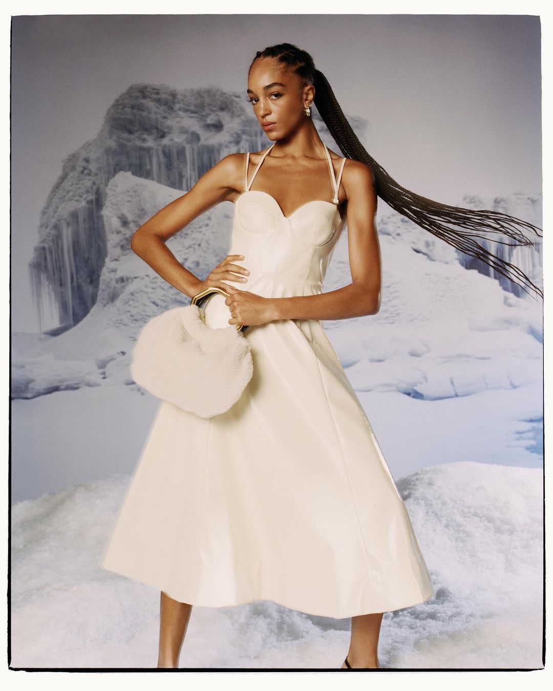Fashion shot of a woman in a cream dress with an oversized faux fur purse, posing in front of a blue and white snowy mountain background.