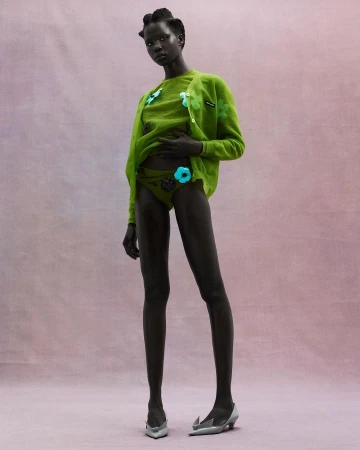 Model wearing a vibrant green cardigan and matching bottoms with floral accents against a textured pink background.